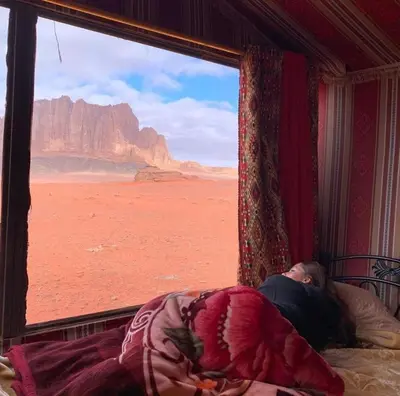 a man laying in bed looking out of a window at the desert at Wadirum sunset camp in Wadi Rum