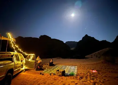 a woman sitting next to a truck on a beach at night at Wadi Rum sleeping in the desert in Wadi Rum