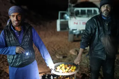 two men standing next to a grill with a basket of fruit at Bedouin Traditions Camp in Wadi Rum