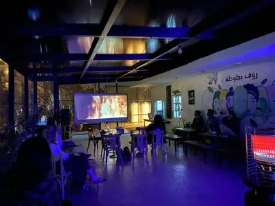 a group of people sitting at tables in a room with a large screen at Battuta Hostel in Amman