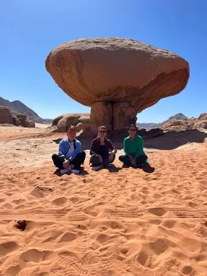 three people sitting in the sand under a rock at The sunset of Wadi Rum in Wadi Rum